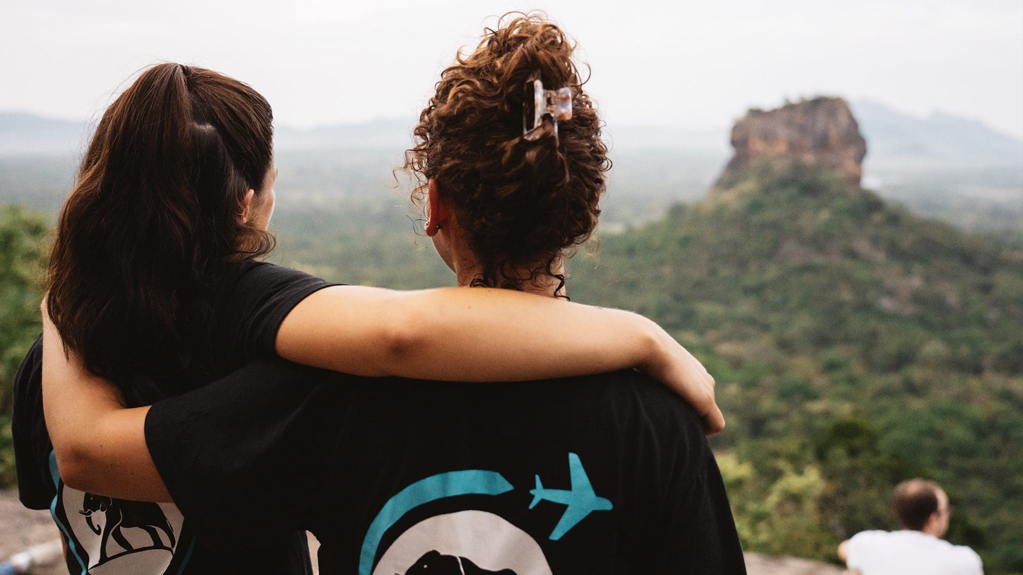 Friends enjoying scenic mountain view beneath moody skies, wearing travel-themed clothing in lush nature.