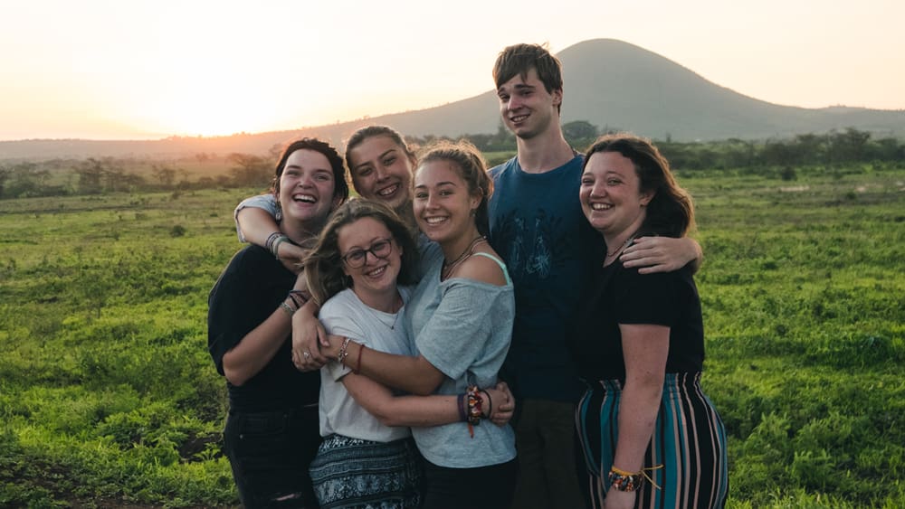 Six friends cheer at sunset in lush green hills, showcasing friendship and outdoor adventure.