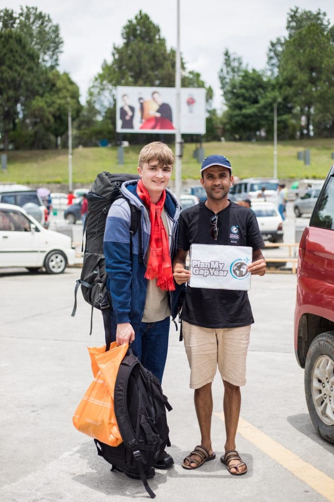 Travelers greeting a local at busy parking lot near transportation hub with parked cars and billboards.